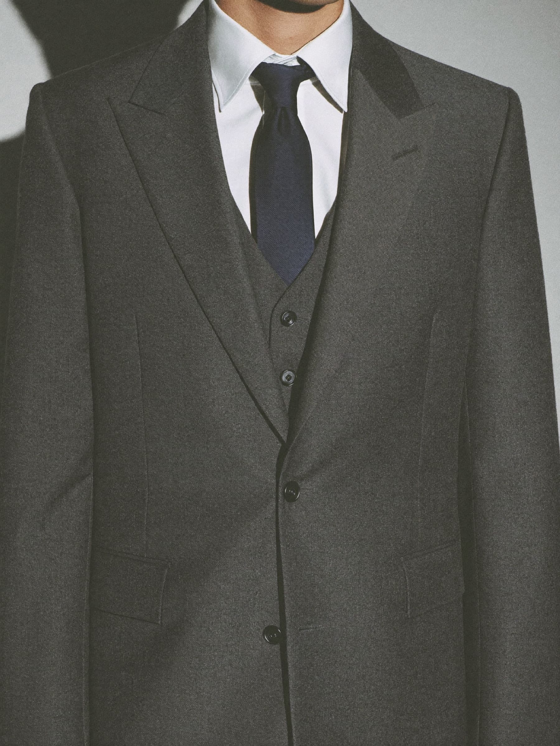Buste d'un homme en costume de laine gris anthracite, chemise blanche et cravate bleu nuit, sur fond de béton clair